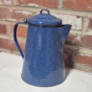 May include: A blue enamel coffee pot with a speckled white finish, featuring a tapered cylindrical body, a hinged lid with a small handle, a curved side handle, and a wide pouring spout. The surface has a glossy, mottled appearance.