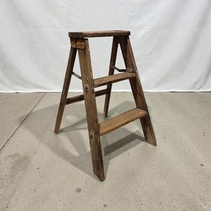 May include: A vintage wooden step stool with three steps. The stool is made of weathered wood, showing signs of age and use. The design features a sturdy A-frame structure with a top step and two lower steps for climbing. The wood has a natural, aged finish.