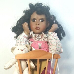 May include: A porcelain doll with dark brown curly hair, wearing a pink and white floral dress with a pink overall, holding a white teddy bear. The doll is sitting on a wooden chair.