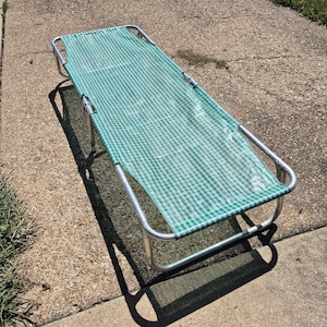 May include: A portable folding cot featuring a rectangular metal frame with a green and white checkered fabric surface. The lightweight aluminum structure includes four legs and horizontal support bars, designed for flat, elevated resting.