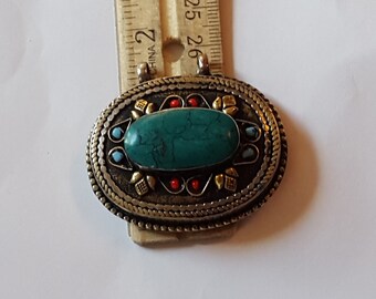 May include: An oval-shaped pendant with a turquoise stone set in a silver and gold frame. The frame is decorated with small red and blue stones.