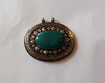 May include: A silver and gold pendant with a turquoise stone set in the center. The pendant is oval shaped and has a detailed design around the stone.