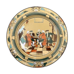 May include: A round ceramic plate with a brown rim and a green and white border. The center of the plate features a scene of five men sitting around a table in a room. The text "An Evening at Ye Lion Inn" is printed at the bottom of the plate.