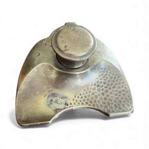 May include: Antique brass inkwell with a round top and a textured, hammered surface. The inkwell has a curved, asymmetrical shape and a small, circular lid. The brass has a tarnished, aged appearance, suggesting its vintage. A classic desk accessory.