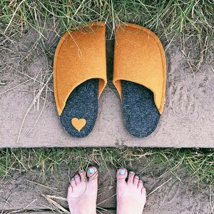 Op de afbeelding: Een paar mosterdgele wollen pantoffels met een donkergrijze wollen voering en een klein hartdetail op de teen. De pantoffels liggen op een houten oppervlak met gras op de achtergrond.