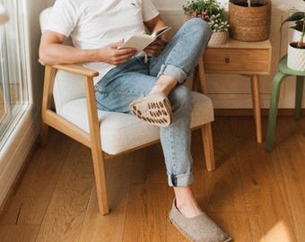 Può includere: Una persona con pantofole grigio chiaro che legge un libro mentre è seduta su una sedia di legno. Le pantofole sono di colore neutro e hanno un design strutturato. La persona indossa una maglietta bianca e jeans blu.