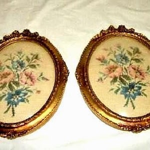 May include: Two antique oval framed needlepoint artworks. Each features a floral bouquet in pink, blue, and green on a cream background. The frames are ornate, with a gold finish and decorative detailing, adding a vintage aesthetic.