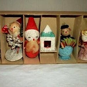 May include: A vintage Christmas ornament set in a cardboard box. The set includes a snowman, a doll, a Santa Claus figure, a house, a clown, and an angel. The ornaments are colorful and detailed, with a retro aesthetic.