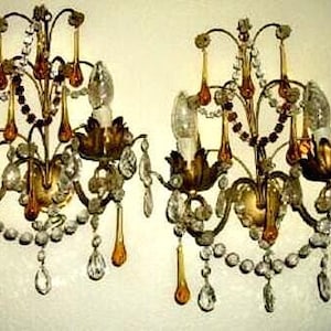 May include: Two ornate wall sconces with gold-toned metal frames, adorned with clear and amber-colored crystal accents. Each sconce features a floral design and two light bulbs. The sconces are mounted on a light-colored wall.