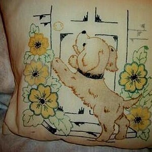 May include: Cream-colored pillow with an embroidered design. The design features a light brown dog reaching for a round object near a black and white door. Yellow flowers with green leaves are on the pillow.