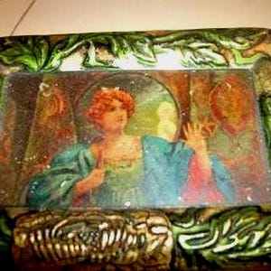May include: An antique rectangular box with a painted image of a woman in a blue gown. The box has a green and brown decorative border with leaf and scroll designs. The bottom of the box has a textured design.