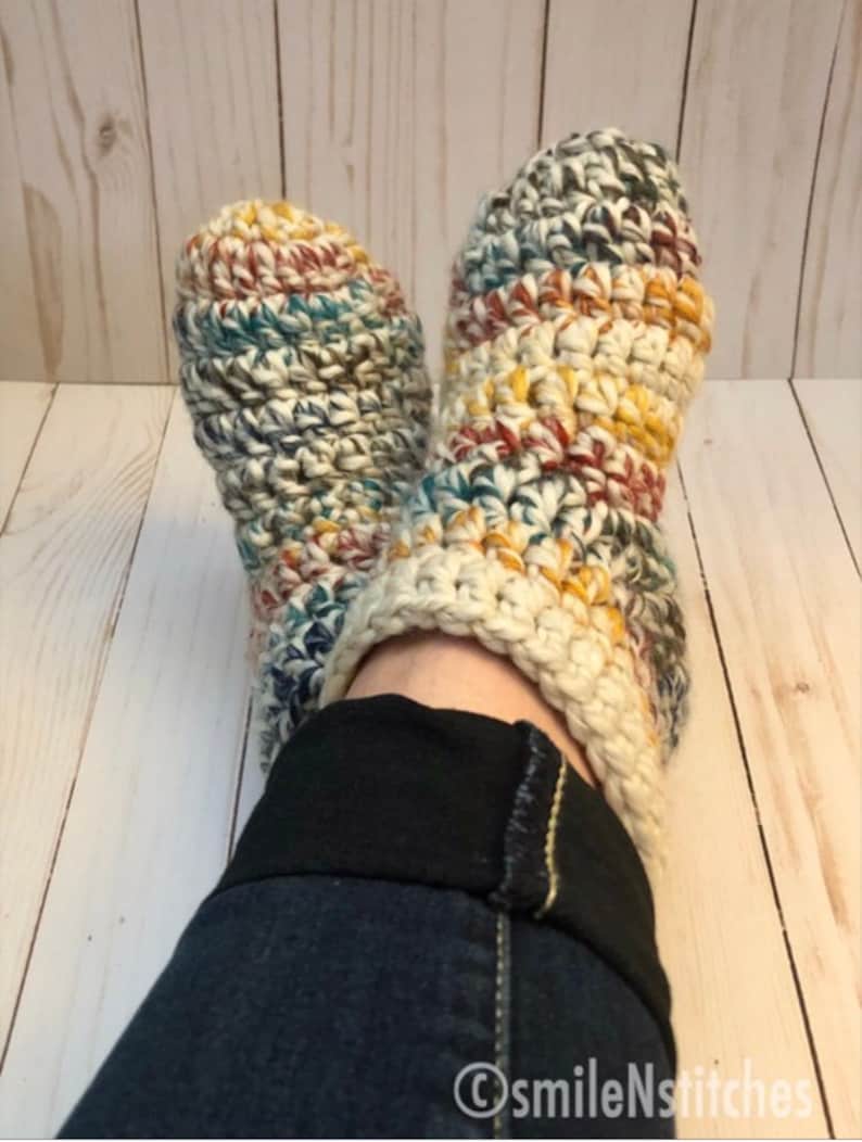 May include: A pair of crocheted slippers with a multi-colored yarn in shades of brown, yellow, blue, and white. The slippers are worn on a person's feet with the bottom of the slippers resting on a wooden surface.