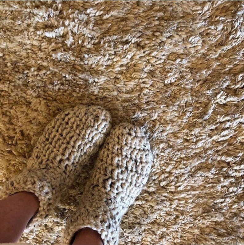 May include: A pair of cozy, knitted beige slippers with a fluffy texture. The slippers are worn on a shaggy, beige rug.
