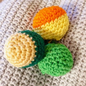 May include: Three crocheted spheres in bright colours: orange and yellow, green, and green and cream. The handmade spheres are arranged on a textured, light grey surface. The spheres appear to be toys or decorative items.