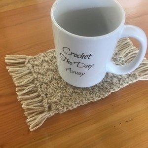 May include: A white ceramic mug with the text "Crochet the Day Away" on it, sitting on a crocheted coaster with fringe.