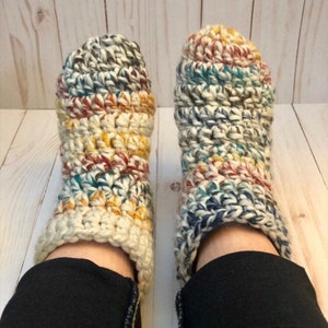 May include: A pair of crocheted slippers with a multi-colored yarn pattern. The slippers are white with blue, red, yellow, and green accents. The slippers are worn on a person's feet.