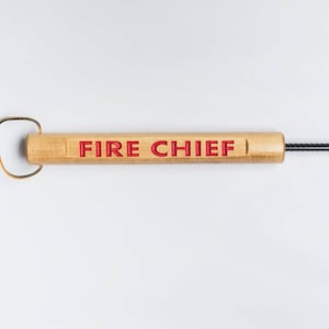 May include: A wooden fire poker with the words "FIRE CHIEF" in red lettering. The poker has a light brown wooden handle with a leather loop and a black metal shaft. The background is white.