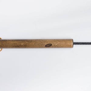Puede incluir: Una varilla de madera con un alambre de metal negro unido a un extremo. La varilla de madera tiene un lazo de cuero marr&oacute;n unido al otro extremo.