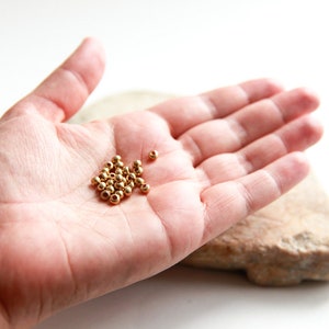 Peut inclure: Un ensemble de petites perles rondes de couleur or. Les perles sont regroupées dans la paume d'une main, avec une pierre de ton neutre en arrière-plan. Les perles semblent être en métal.