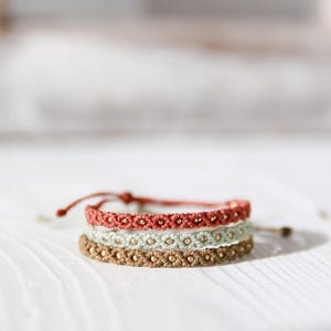 Peut inclure: Trois bracelets en macramé, rouge, vert et marron, ornés de petites perles dorées. Ils sont empilés sur une surface en bois blanc, mettant en valeur leur motif tissé complexe et leurs tons naturels.