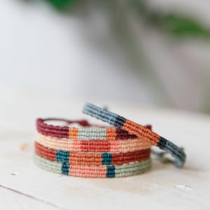 Peut inclure: Plusieurs bracelets tissés colorés. Les bracelets présentent un motif répétitif de couleurs variées, notamment rouge, orange, turquoise et gris. Un bracelet est légèrement séparé de la pile. Les bracelets sont sur une surface en bois blanc.
