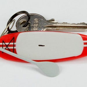 K&ouml;nnte beinhalten: Ein Miniatur-Schl&uuml;sselanh&auml;nger in Form eines rot-wei&szlig;en Stand-Up-Paddleboards mit einem kleinen grauen Paddel, das an einem Drahtseil befestigt ist. Das Board hat ein wei&szlig;es Deckpad, rote Kanten mit wei&szlig;en Streifen und ein schwarzes Grafikdetail.