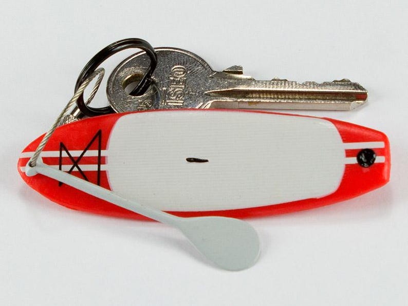 K&ouml;nnte beinhalten: Ein Miniatur-Schl&uuml;sselanh&auml;nger in Form eines rot-wei&szlig;en Stand-Up-Paddleboards mit einem kleinen grauen Paddel, das an einem Drahtseil befestigt ist. Das Board hat ein wei&szlig;es Deckpad, rote Kanten mit wei&szlig;en Streifen und ein schwarzes Grafikdetail.