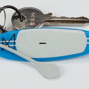 K&ouml;nnte beinhalten: Ein blau-wei&szlig;er Miniatur-Stand-Up-Paddleboard-Schl&uuml;sselanh&auml;nger mit einem kleinen grauen Paddel, das an einer Drahtschlaufe befestigt ist. Das Board hat ein wei&szlig;es Deckpad, blaue Kanten mit wei&szlig;en Streifen und einen schwarzen Schl&uuml;sselring.