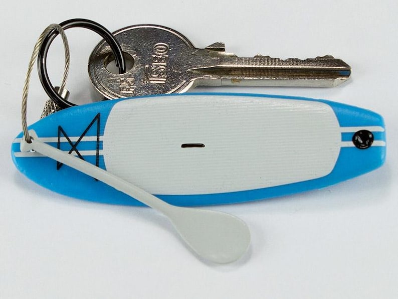 K&ouml;nnte beinhalten: Ein blau-wei&szlig;er Miniatur-Stand-Up-Paddleboard-Schl&uuml;sselanh&auml;nger mit einem kleinen grauen Paddel, das an einer Drahtschlaufe befestigt ist. Das Board hat ein wei&szlig;es Deckpad, blaue Kanten mit wei&szlig;en Streifen und einen schwarzen Schl&uuml;sselring.