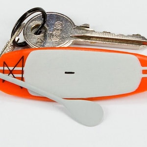 K&ouml;nnte beinhalten: Ein Miniatur-Schl&uuml;sselanh&auml;nger in Form eines orange-wei&szlig;en Stand-Up-Paddleboards mit einem kleinen grauen Paddel an einem Draht sowie einem schwarzen Metallring mit einem silberfarbenen ISEO-Schl&uuml;ssel. Das Board hat ein wei&szlig;es Deckpad und ein schwarzes Logo.