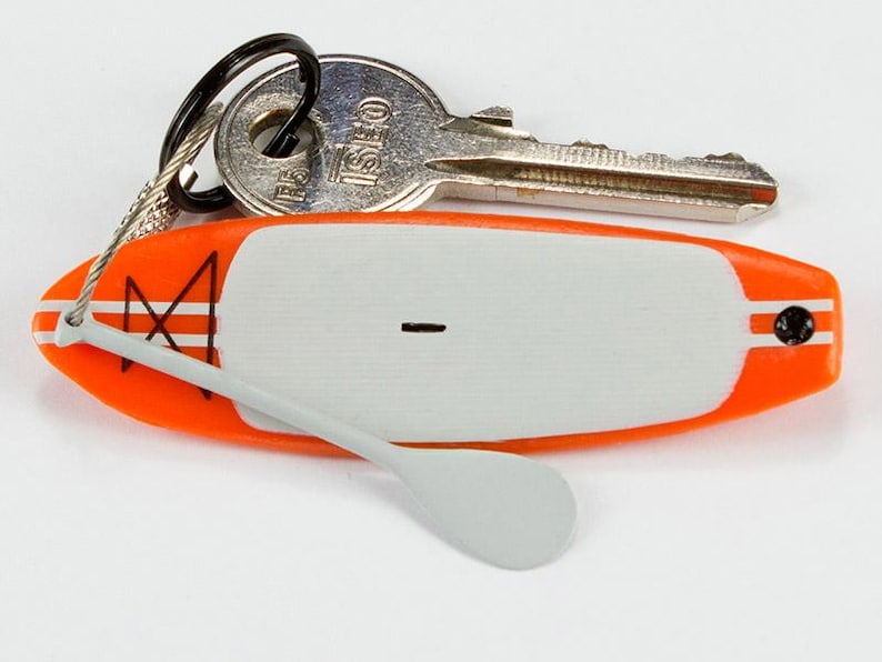 K&ouml;nnte beinhalten: Ein Miniatur-Schl&uuml;sselanh&auml;nger in Form eines orange-wei&szlig;en Stand-Up-Paddleboards mit einem kleinen grauen Paddel an einem Draht sowie einem schwarzen Metallring mit einem silberfarbenen ISEO-Schl&uuml;ssel. Das Board hat ein wei&szlig;es Deckpad und ein schwarzes Logo.
