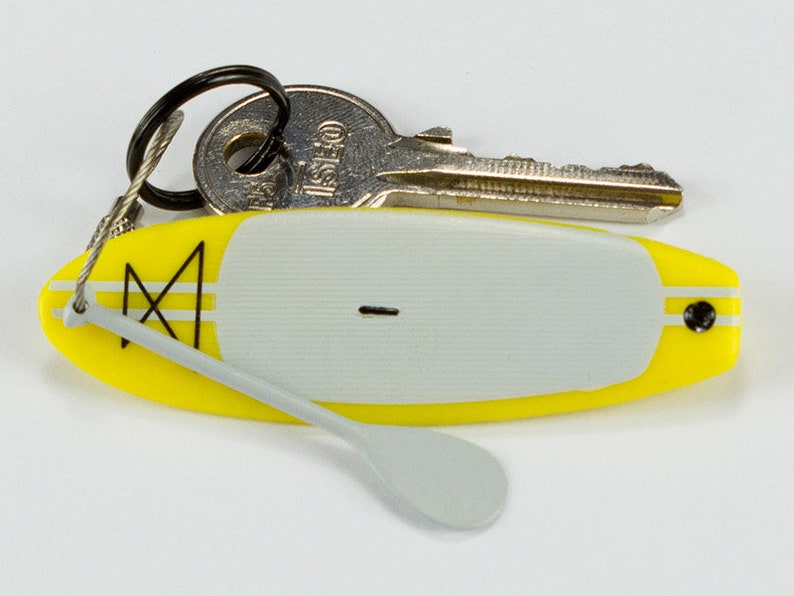 K&ouml;nnte beinhalten: Ein gelb-grauer Miniatur-Surfbrett-Schl&uuml;sselanh&auml;nger mit einem kleinen grauen Paddel, das an einem Drahtseil befestigt ist. Ein silberner Metallschl&uuml;ssel h&auml;ngt ebenfalls am schwarzen Ring. Das Surfbrett hat ein graues Deckpad und ein schwarzes Logo.