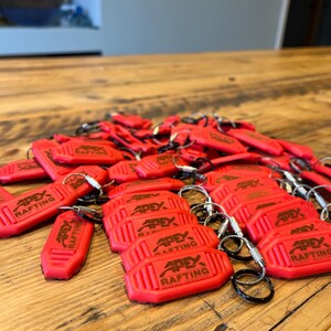 K&ouml;nnte beinhalten: Eine Ansammlung rechteckiger roter Kunststoff-Schl&uuml;sselanh&auml;nger, jeweils mit einem Metallring und dem gepr&auml;gten Text APEX RAFTING versehen. Die Schl&uuml;sselanh&auml;nger haben eine strukturierte Oberfl&auml;che mit vertikalen Rillen an den Seiten.
