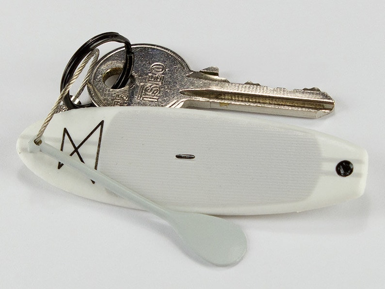 K&ouml;nnte beinhalten: Ein wei&szlig;er Miniatur-Surfbrett-Schl&uuml;sselanh&auml;nger mit einem schwarzen geometrischen Logo und einem kleinen befestigten wei&szlig;en Paddel. Das Surfbrett ist an einem silbernen Metallring befestigt, der einen silbernen ISEO-Schl&uuml;ssel mit schwarzem Ring h&auml;lt.