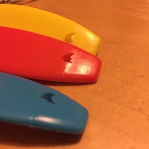 K&ouml;nnte beinhalten: Drei kleine, surfboardf&ouml;rmige Objekte in den leuchtenden Farben Gelb, Rot und Blau. Jedes St&uuml;ck hat eine glatte, gew&ouml;lbte Oberfl&auml;che mit einem kleinen, erhabenen Flossendetail in der N&auml;he des abgerundeten Endes und eine gl&auml;nzende Oberfl&auml;che.