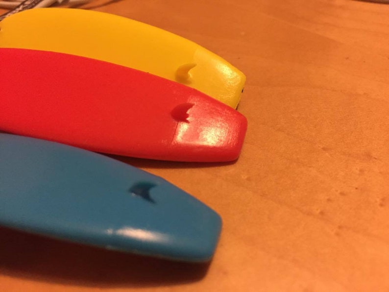 K&ouml;nnte beinhalten: Drei kleine, surfboardf&ouml;rmige Objekte in den leuchtenden Farben Gelb, Rot und Blau. Jedes St&uuml;ck hat eine glatte, gew&ouml;lbte Oberfl&auml;che mit einem kleinen, erhabenen Flossendetail in der N&auml;he des abgerundeten Endes und eine gl&auml;nzende Oberfl&auml;che.