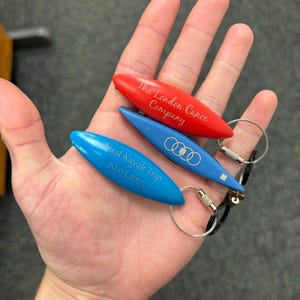 K&ouml;nnte beinhalten: Drei kanuf&ouml;rmige Schl&uuml;sselanh&auml;nger in Rot und Blau mit eingravierten Texten und Logos. Der rote Anh&auml;nger tr&auml;gt die Aufschrift The London Canoe Company. Die blauen Anh&auml;nger zeigen das Audi-Logo und den Text First Kayak Trip 20.05.2025 an Metallringen.
