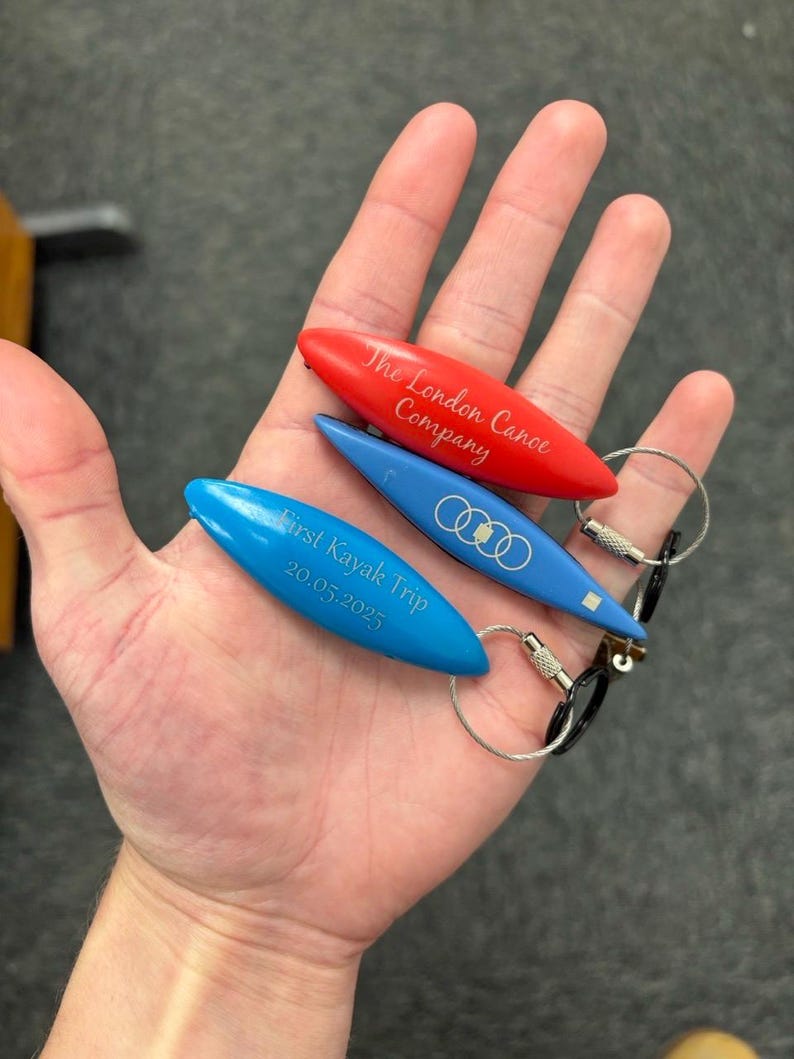 K&ouml;nnte beinhalten: Drei kanuf&ouml;rmige Schl&uuml;sselanh&auml;nger in Rot und Blau mit eingravierten Texten und Logos. Der rote Anh&auml;nger tr&auml;gt die Aufschrift The London Canoe Company. Die blauen Anh&auml;nger zeigen das Audi-Logo und den Text First Kayak Trip 20.05.2025 an Metallringen.