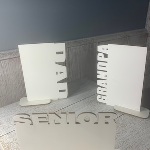 May include: Three white wooden signs with cut-out lettering. The signs read "Senior", "Dad", and "Grandpa".