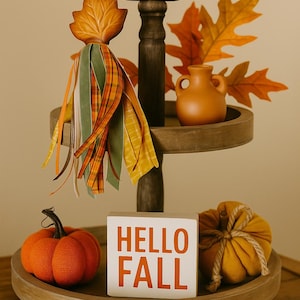 May include: A two-tiered wooden tray decorated for fall. The top tier holds a small orange vase and faux fall leaves. The bottom tier features an orange pumpkin, a fabric pumpkin, and a white block with "HELLO FALL" in orange letters. A leaf decoration hangs from the top.