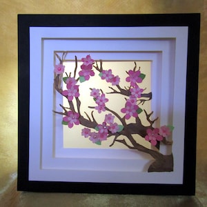 May include: A framed paper art piece featuring a cherry blossom branch with pink flowers and green leaves. The artwork is set against a gold background within a white and black layered frame.