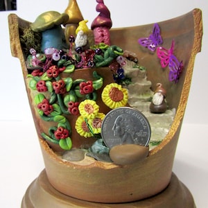 May include: A broken terracotta flower pot with a miniature fairy garden scene inside. The scene includes a small house, a gnome, a butterfly, and a variety of flowers and plants. A US quarter is included for scale.