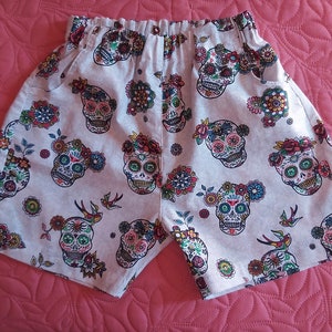 Peut inclure: Shorts de couleur claire avec un imprimé intégral de crânes en sucre colorés décorés de motifs floraux et de petits oiseaux. Le short possède une taille élastique et des poches latérales, présentant un motif répétitif de crânes dans des tons de rouge, bleu, vert et jaune.