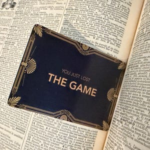 May include: A rectangular dark blue bookmark with gold-colored art deco corner accents and borders. The text "YOU JUST LOST THE GAME" is printed in gold-colored capital letters in the center. The bookmark is placed on an open dictionary page.