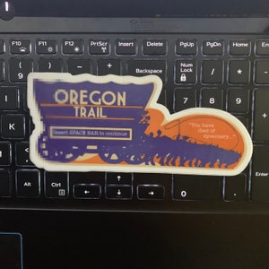 May include: A sticker of a vintage illustration depicting a covered wagon train on the Oregon Trail. The text "Oregon Trail" is in a purple banner above the wagon train. The text "Insert SPACE BAR to continue" is below the wagon train. The text "You have died of dysentery..." is in a speech bubble above the wagon train.