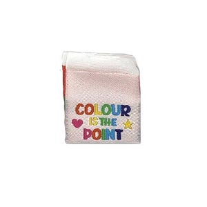 May include: A small, folded fabric label with the embroidered text "COLOUR IS THE POINT" in rainbow colours. A pink heart and yellow star are also embroidered on the label. The label is primarily light pink with white and red accents.