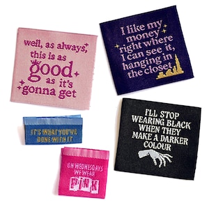May include: A selection of embroidered fabric labels in different colours, each featuring text. The labels display phrases such as "good as it's gonna get," "I like my money," and "I'll stop wearing black." The labels are square and rectangular.