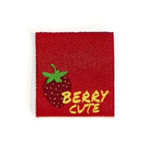 May include: A red square fabric label featuring a strawberry illustration and the words "BERRY CUTE" in yellow. The strawberry has green leaves. The label has a textured appearance.