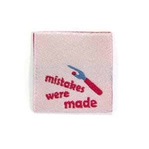 May include: A small, square, pink fabric label with the words "mistakes were made" in red, stylized text. A blue and red graphic of a tool is above the word "made". The label has a white border.