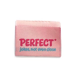 May include: A pink fabric label with the word "PERFECT" embroidered in a bold, magenta font. Below, the phrase "jokes, not even close" is embroidered in a teal colour. The label has a rectangular shape.